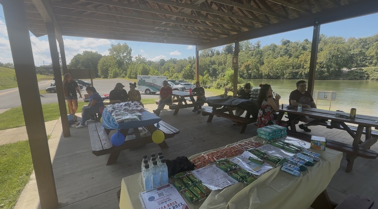 Neighbors at a riverside pavilion with tables of donated food and essentials